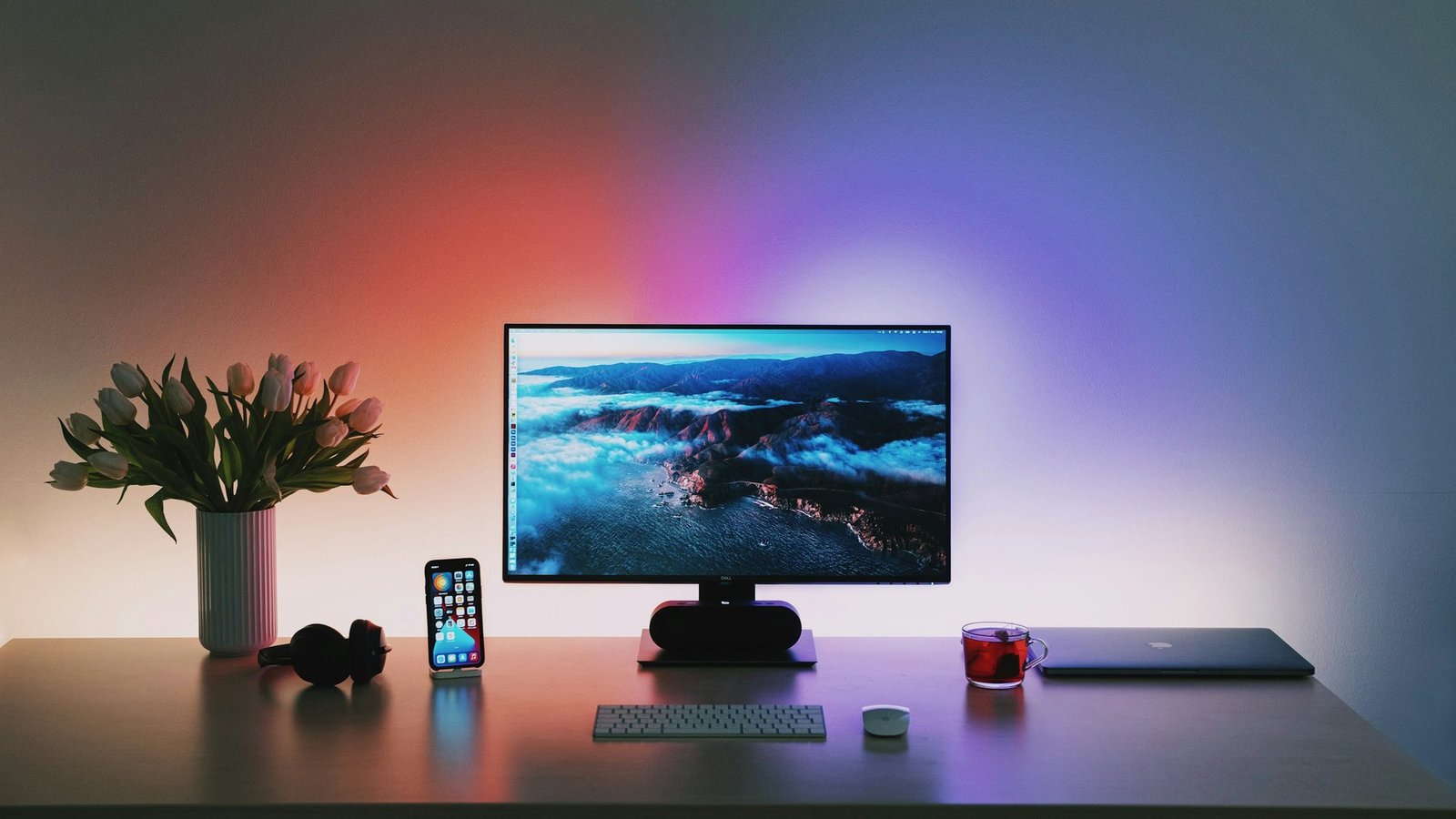 Minimal desk setup with a monitor, keyboard, mouse, headphones, and laptop under purple and pink underglow